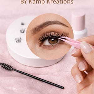 以下が含まれることがあります： 「LASH CLUSTER GUIDE」と「BY Kamp Kreations」の文字が入った、まつげクラスターを宣伝する画像。ピンクのピンセットで長いまつげを装着している目が写っています。まつげクラスターが入った白いトレイ、まつげブラシ、ピンクのボトルも写っています。