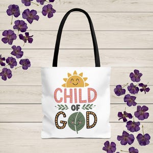 May include: White tote bag with black handles. The bag has a graphic design of a smiling sun with the text "CHILD OF GOD" in pink and yellow letters with a green leaf with a smiling face.