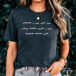 May include: Dark gray t-shirt with white Morse code design. The shirt has a crew neck and short sleeves. The text "Scripture Sayings" is printed in cursive on the lower right side of the shirt.