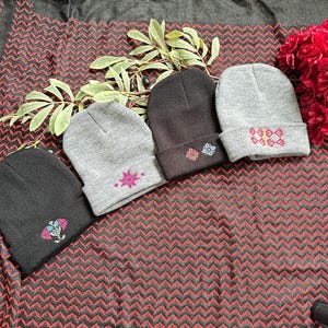 May include: Four beanies in shades of gray and black, each with embroidered designs. The beanies are arranged in a row, showcasing different patterns in pink, blue, and white. The background features a patterned fabric with red and black zigzags.