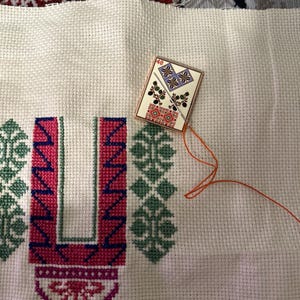 May include: Close-up of a cross-stitch project in progress. The fabric is off-white, with a partially completed design in pink, green, and blue thread. A small, decorative needle minder with a geometric pattern is attached.