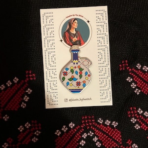 May include: A white ceramic bottle with blue and red floral designs. The bottle is a pin with a gold finish. The pin is on a white card with the text "Falastin By The Stitch" and a social media handle.