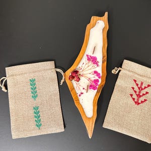 May include: A wooden candle shaped like a map, filled with white wax and dried flowers, is positioned between two burlap drawstring bags. One bag has a green leaf design, the other a red design.