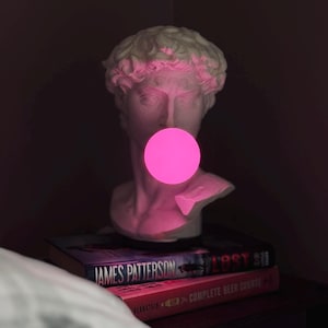 Peut inclure: Un buste en plâtre blanc d'un homme avec une ampoule rose dans la bouche. Le buste est posé sur une pile de livres.