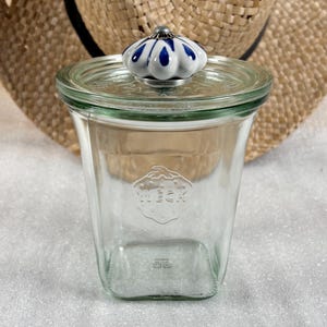 Pode incluir: Um frasco Weck de vidro transparente com tampa e um botão decorativo azul e branco. O frasco tem uma base quadrada e um corpo ligeiramente alargado. O logotipo Weck está em relevo na frente. Ideal para conservação de alimentos.