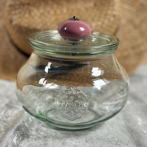 Może przedstawiać: Przezroczysty szklany słoik z pokrywką, z różowym guzikiem na górze. Słoik ma zaokrąglony kształt i widoczne wytłoczone logo. Słoik stoi na białej powierzchni.