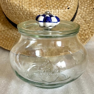 Może przedstawiać: Przezroczysty szklany słoik z pokrywką i niebiesko-białym, szachownicowym pokrętłem. Słoik ma zaokrąglony kształt, a na przodzie wytłoczono napis "WECK". W tle znajduje się słomkowy kapelusz.