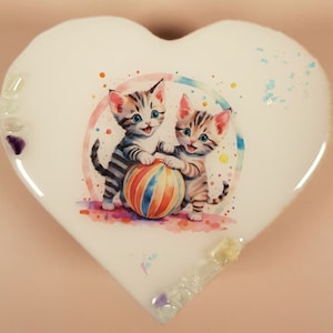 Könnte beinhalten: Weiße herzförmige Keramik-Schmuckschatulle mit einer bunten Illustration von zwei Kätzchen, die mit einem gestreiften Ball spielen. Die Schachtel ist mit schimmerndem Glitzer verziert.