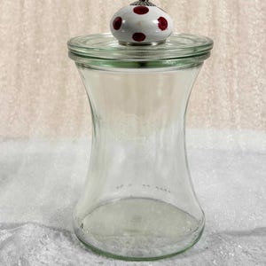 Puede incluir: Un tarro de cristal transparente con una tapa decorativa. La tapa es blanca con lunares rojos y una base plateada. El tarro tiene una base ancha que se estrecha hacia adentro y luego se ensancha hacia arriba. El tarro está vacío.