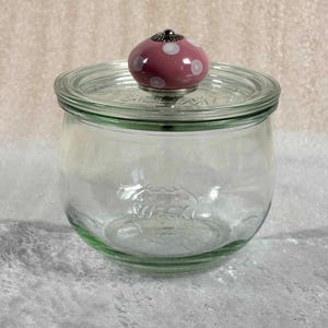 Op de afbeelding: Een heldere glazen pot met een deksel en een roze knop met witte stippen. De pot heeft een brede opening en een afgeronde body. Het deksel is van glas en heeft een metalen rand. De pot staat op een wit oppervlak.