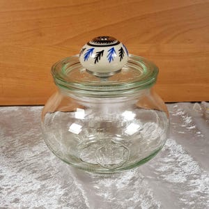 Könnte beinhalten: Ein klares Glasgefäß mit einem weißen Keramikdeckel mit einem blauen und schwarzen Blumendesign. Das Gefäß hat eine breite Öffnung und einen abgerundeten Körper.