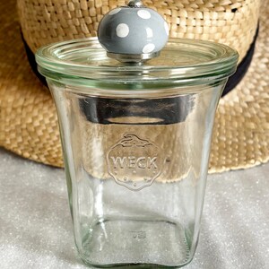 Puede incluir: Un tarro de cristal transparente con tapa y pomo gris con lunares blancos. El tarro tiene la palabra "WECK" grabada en relieve en la parte delantera. El tarro está delante de un sombrero de paja.