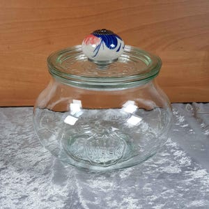Op de afbeelding: Een glazen pot met een brede opening en een keramische deksel met een bloemenpatroon. De pot heeft een reliëf WECK-logo op de bodem.