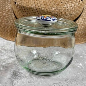 Może przedstawiać: Przezroczysty szklany słoik z pokrywką i dekoracyjnym guzikiem z niebieskim, pomarańczowym i białym wzorem. Słoik ma zaokrąglony kształt i zieloną podstawę. Z przodu wytłoczony jest napis "WECK".