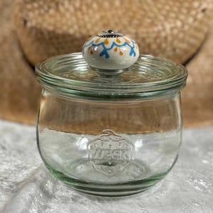 Op de afbeelding: Een heldere glazen pot met een deksel en een decoratieve knop. De knop is wit met blauwe en oranje bloemmotieven. De pot heeft een Weck-logo op de voorkant en staat op een wit oppervlak.