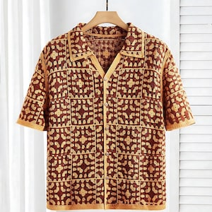 Op de afbeelding: Een bruin en beige gehaakt overhemd met korte mouwen en een knoopsluiting. Het overhemd heeft een kraag en een vierkant patroon. Het gehaakte overhemd hangt aan een houten hanger tegen een witte achtergrond.