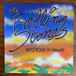 May include: A vintage concert poster for The Rolling Stones' 1973 tour in Hawaii. The poster features the band's name in a stylized script with a black outline. The background has a gradient of blue, yellow, orange, and green, with palm tree silhouettes.