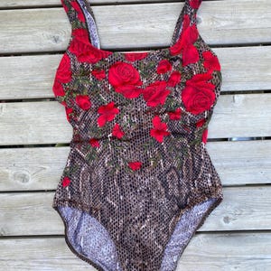May include: One-piece swimsuit with a rose and snakeskin pattern. The top features large red roses and green leaves on a black background. The bottom has a brown snakeskin print. The swimsuit has wide straps.