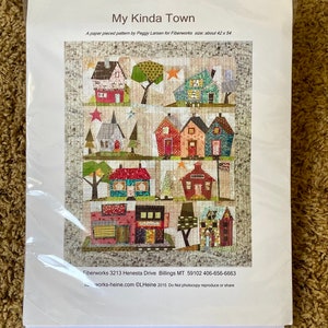 May include: A paper pieced quilt pattern titled "My Kinda Town" by Peggy Larsen for Fiberworks. The pattern features a colorful patchwork design of houses and trees. The size is about 42 x 54 inches.