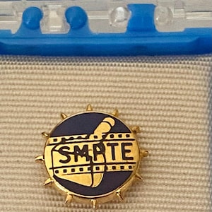 May include: A gold-colored pin with a navy blue center featuring the text "SMPTE" in gold. The pin has a circular design with a film strip and a stylized image. The pin is surrounded by gold-colored spikes.