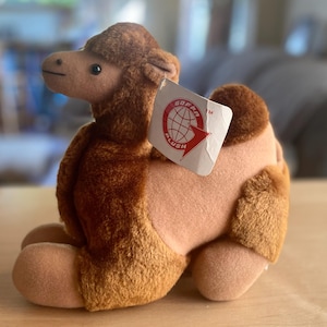 May include: A plush camel toy in shades of brown and tan, with a tag that reads "GOFRA PLUSH". The stuffed animal is sitting on a wooden surface. The camel has a detailed face and is made of soft material.
