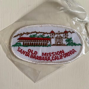 May include: An oval-shaped patch featuring an embroidered image of the Old Mission Santa Barbara, California. The patch has a white background with a detailed depiction of the mission in brown and red, with green palm trees and text that reads "Old Mission Santa Barbara, California."
