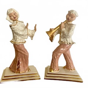 May include: A pair of ceramic figurines depicting musicians. One figure is singing, the other playing a gold trumpet. Both figures wear white tops with gold trim and pink pants. The figurines stand on cream-colored bases with gold accents.