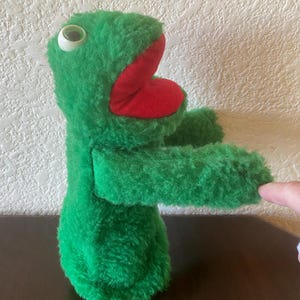 May include: A green plush puppet with a red mouth and large white eyes. The puppet has a fuzzy texture and is designed to be worn on the hand. The puppet's arms are outstretched, and it appears to be a whimsical character.