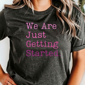 May include: Dark gray t-shirt with the text "We Are Just Getting Started" in pink. The shirt has short sleeves and a slightly faded appearance, suggesting a vintage or worn look. The overall style is casual and comfortable.