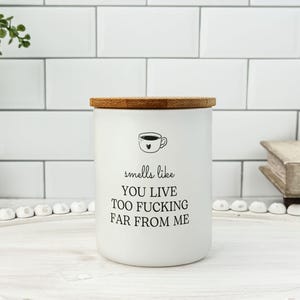 K&ouml;nnte beinhalten: Wei&szlig;es Keramik-Kerzenglas mit Bambusdeckel. Das Glas hat eine schwarze Grafik einer Kaffeetasse und den Text "smells like you live too fucking far from me."