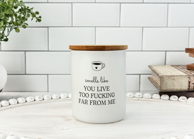 K&ouml;nnte beinhalten: Wei&szlig;es Keramik-Kerzenglas mit Bambusdeckel. Das Glas hat eine schwarze Grafik einer Kaffeetasse und den Text "smells like you live too fucking far from me."