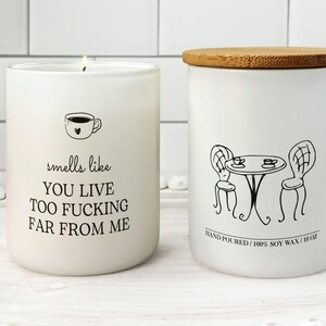 K&ouml;nnte beinhalten: Zwei wei&szlig;e Sojawachskerzen. Eine Kerze hat eine Kaffeetassen-Grafik und den Text "smells like YOU LIVE TOO FUCKING FAR FROM ME." Die andere Kerze hat eine schwarze Strichzeichnung eines Tisches und St&uuml;hlen. Die Kerze ist 283 g.