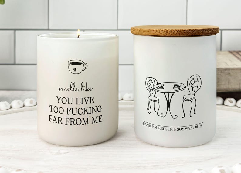 K&ouml;nnte beinhalten: Zwei wei&szlig;e Sojawachskerzen. Eine Kerze hat eine Kaffeetassen-Grafik und den Text "smells like YOU LIVE TOO FUCKING FAR FROM ME." Die andere Kerze hat eine schwarze Strichzeichnung eines Tisches und St&uuml;hlen. Die Kerze ist 283 g.