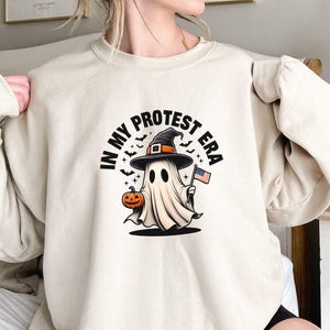 May include: Beige sweatshirt featuring a cartoon ghost wearing a witch hat and holding a pumpkin and an American flag. The text "IN MY PROTEST ERA" is printed above the ghost in a circular design. Bats are also included in the design.