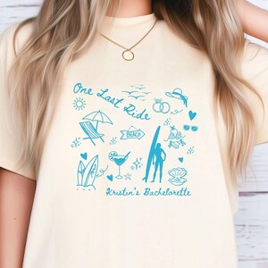 May include: A light yellow t-shirt with a blue graphic design featuring a surfboard, beach umbrella, and the text "One Last Ride" and "Kristin's Bachelorette".