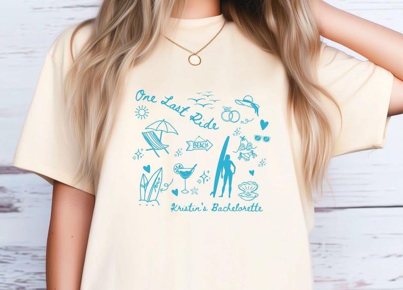 May include: A light yellow t-shirt with a blue graphic design featuring a surfboard, beach umbrella, and the text "One Last Ride" and "Kristin's Bachelorette".