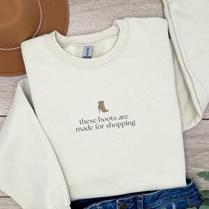 May include: A cream-colored sweatshirt with the phrase "these boots are made for shopping" and a small boot graphic. A brown hat and blue jeans are also in the image.