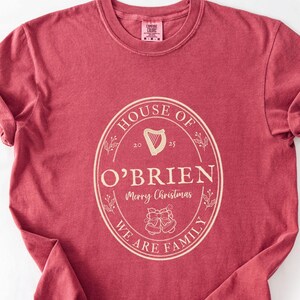 May include: A maroon t-shirt with a cream-colored oval design. The design includes the words "HOUSE OF O'BRIEN" and "WE ARE FAMILY", with a harp and bells graphic. The shirt has rolled-up sleeves.
