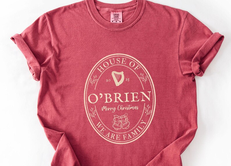 May include: A maroon t-shirt with a cream-colored oval design. The design includes the words "HOUSE OF O'BRIEN" and "WE ARE FAMILY", with a harp and bells graphic. The shirt has rolled-up sleeves.