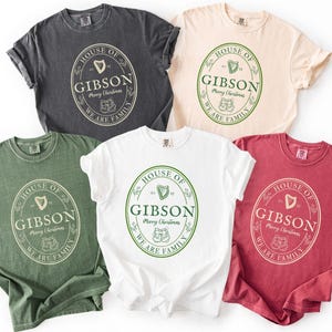 May include: Five t-shirts in various colors: charcoal, cream, green, white, and red. Each shirt features a circular design with the text "HOUSE OF GIBSON Merry Christmas WE ARE FAMILY" in a vintage style. The design includes a harp and two stockings.