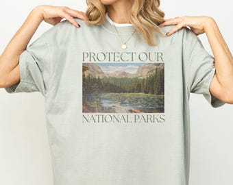 May include: A light green t-shirt with the text "PROTECT OUR NATIONAL PARKS" printed on the front. The shirt features a vintage-style image of a lake surrounded by trees and mountains.