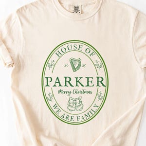May include: Cream-colored t-shirt with a green oval design. The design includes the words "HOUSE OF PARKER", "Merry Christmas", and "WE ARE FAMILY". A harp and bells are also featured.