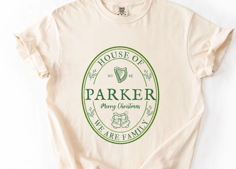 May include: Cream-colored t-shirt with a green oval design. The design includes the words "HOUSE OF PARKER", "Merry Christmas", and "WE ARE FAMILY". A harp and bells are also featured.