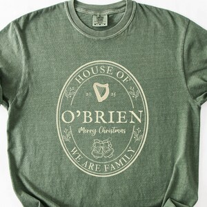May include: Sage green t-shirt with rolled sleeves. The front features a cream-colored oval design with the words "HOUSE OF O'BRIEN" and "WE ARE FAMILY". A harp and the words "Merry Christmas" are also included.