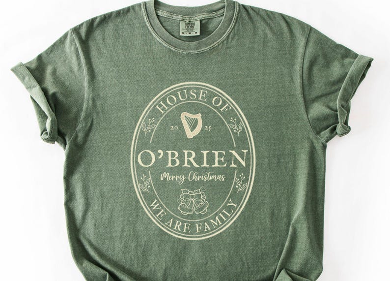May include: Sage green t-shirt with rolled sleeves. The front features a cream-colored oval design with the words "HOUSE OF O'BRIEN" and "WE ARE FAMILY". A harp and the words "Merry Christmas" are also included.