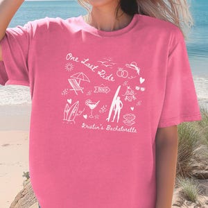 May include: Pink t-shirt with a white graphic design that says "One Last Ride" with beach-themed illustrations, including a surfboard, a beach chair, a cocktail, and the text "Kristin's Bachelorette".