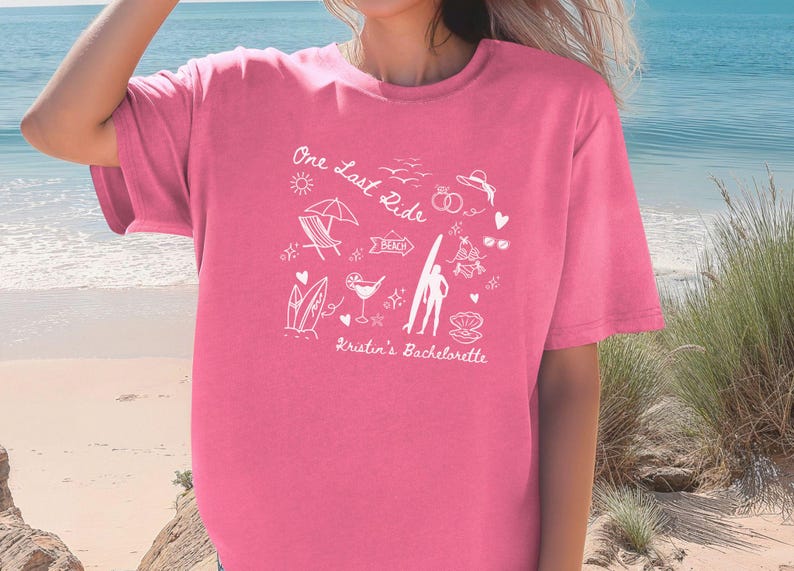 May include: Pink t-shirt with a white graphic design that says "One Last Ride" with beach-themed illustrations, including a surfboard, a beach chair, a cocktail, and the text "Kristin's Bachelorette".