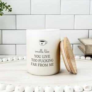 K&ouml;nnte beinhalten: Eine cremefarbene Kerze mit Holzdeckel, mit dem Text "smells like you live too fucking far from me" und einer Kaffeetassen-Grafik. Die Kerze steht auf einem wei&szlig;en Tablett mit dekorativen Perlen.