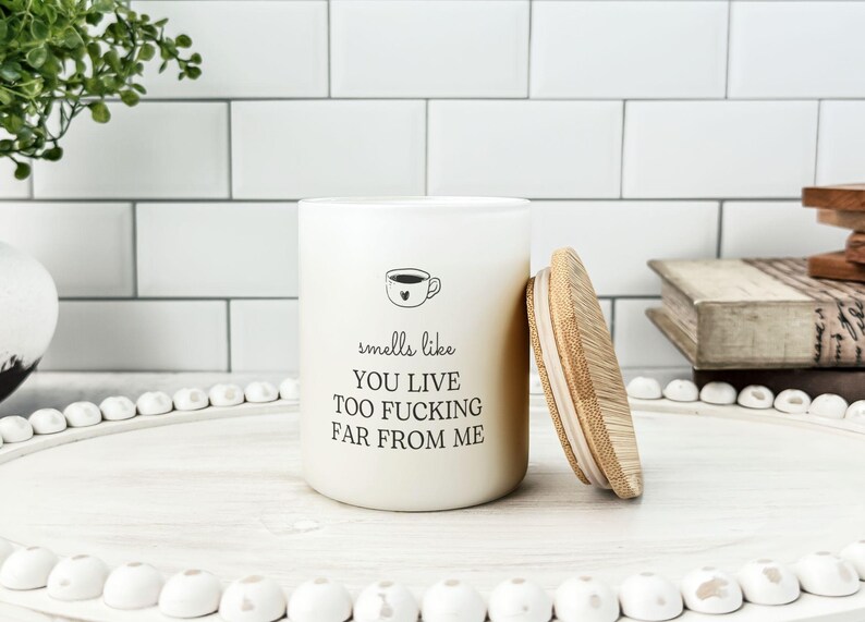 K&ouml;nnte beinhalten: Eine cremefarbene Kerze mit Holzdeckel, mit dem Text "smells like you live too fucking far from me" und einer Kaffeetassen-Grafik. Die Kerze steht auf einem wei&szlig;en Tablett mit dekorativen Perlen.