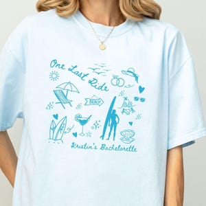 May include: Light blue t-shirt with a blue graphic design of a beach scene with the text "One Last Ride" and "Kristin's Bachelorette".